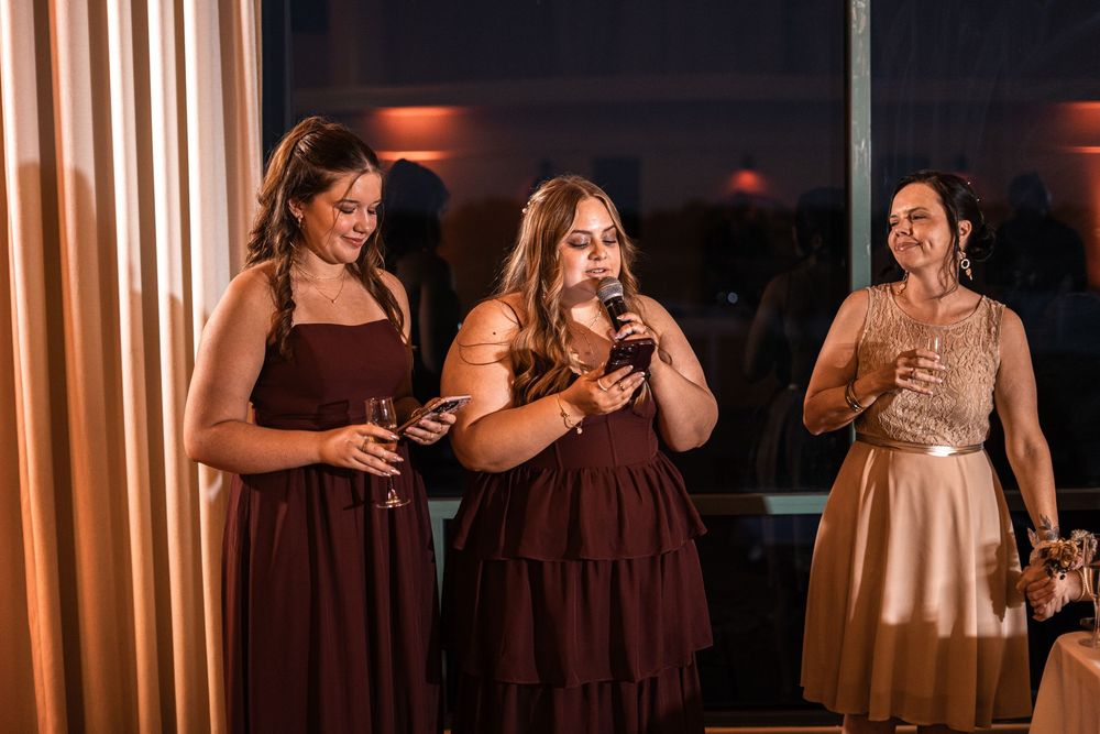 Three bridesmaids in burgundy dresses laughing and holding champagne glasses at a wedding reception.