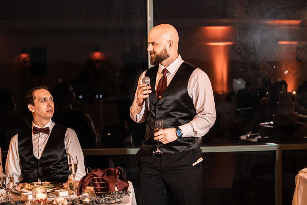 Bald groom with beard delivers wedding speech holding microphone and glass at elegant reception dinner.