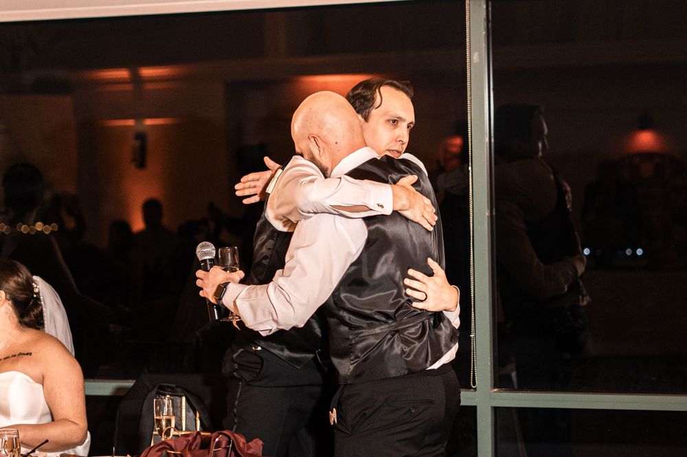 Two men share an emotional hug at a wedding reception, one holding a microphone, with guests seated nearby.