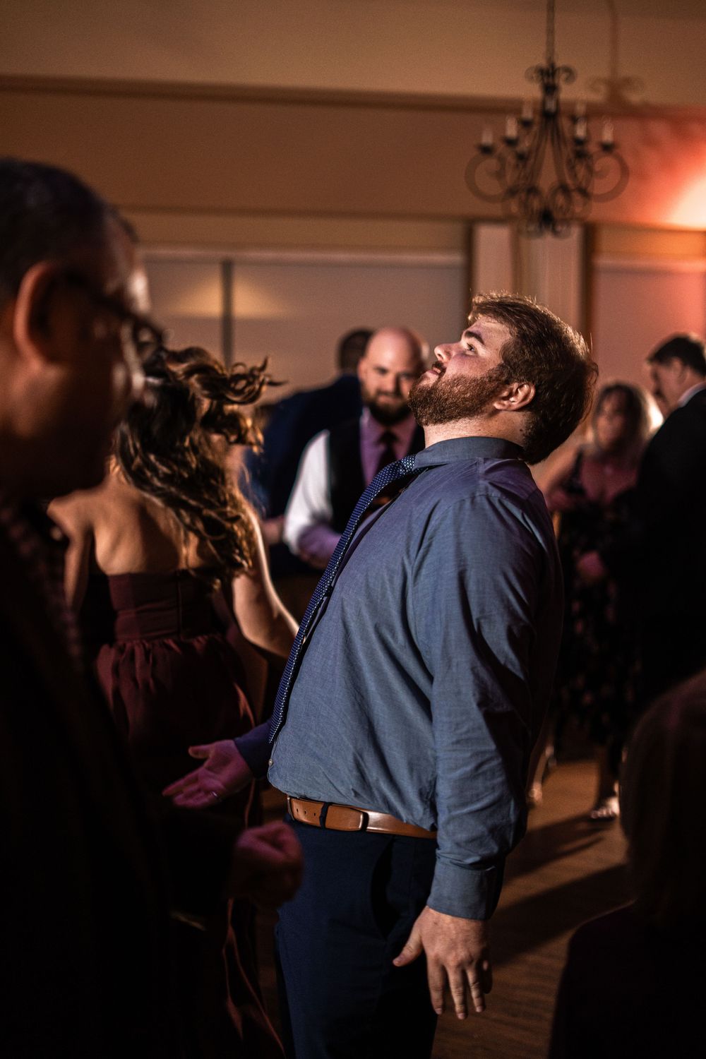 Bearded man in blue shirt dancing joyfully at a wedding reception in a warmly lit ballroom.