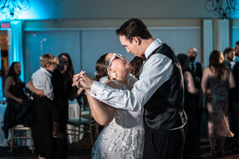 Couple shares romantic dance at wedding reception with blue ambient lighting and guests in background.