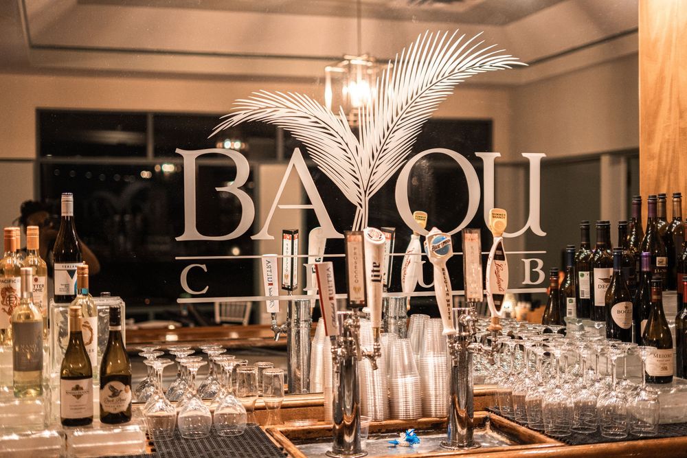 Bayou Club bar display with champagne bottles, glassware, and a decorative palm leaf under warm ambient lighting.