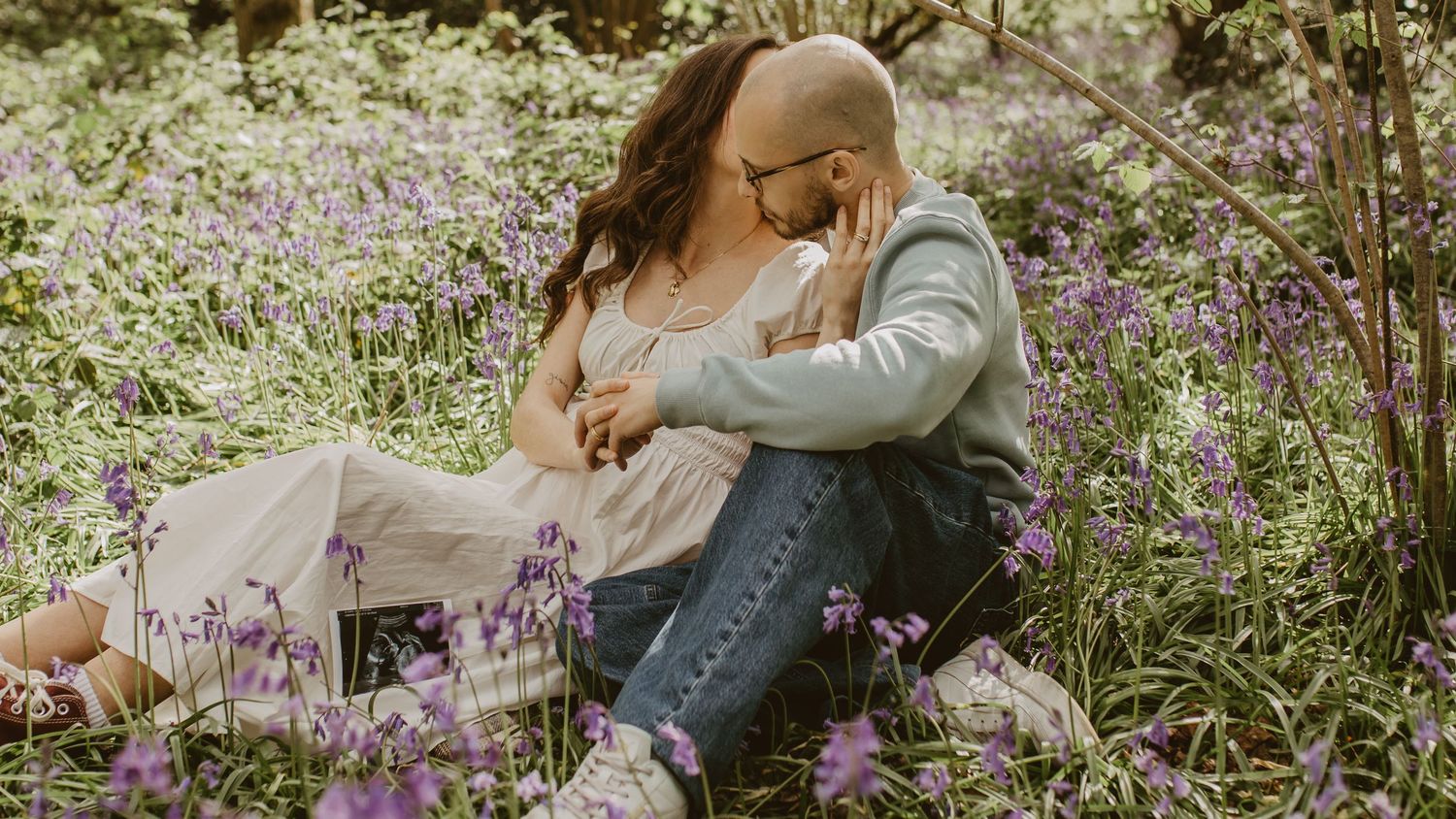 A couple sits romantically among purple wildflowers in a lush green meadow, sharing a tender embrace outdoors.