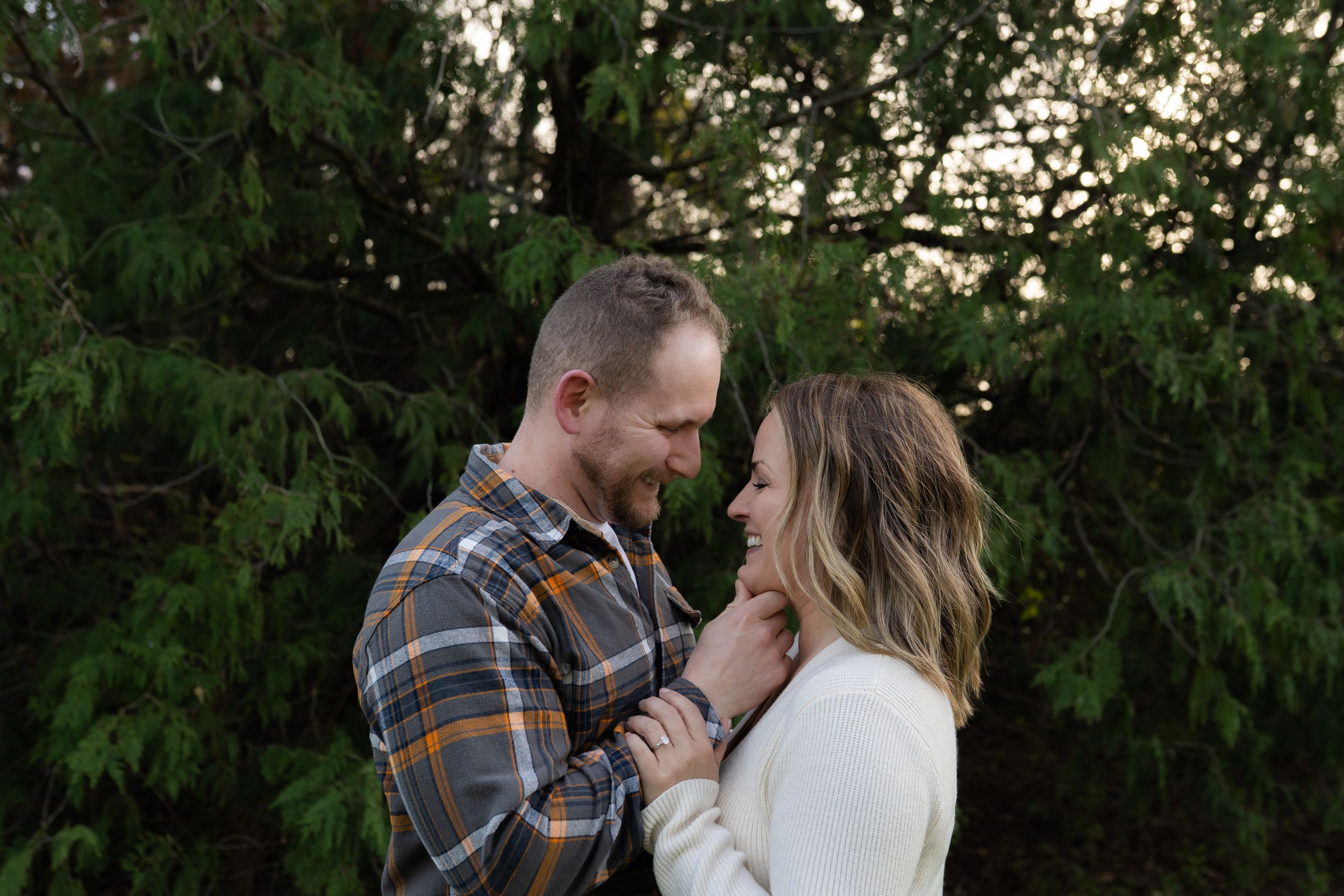 Couple sharing a romantic moment smiling at each other outdoors near evergreen trees at golden hour.