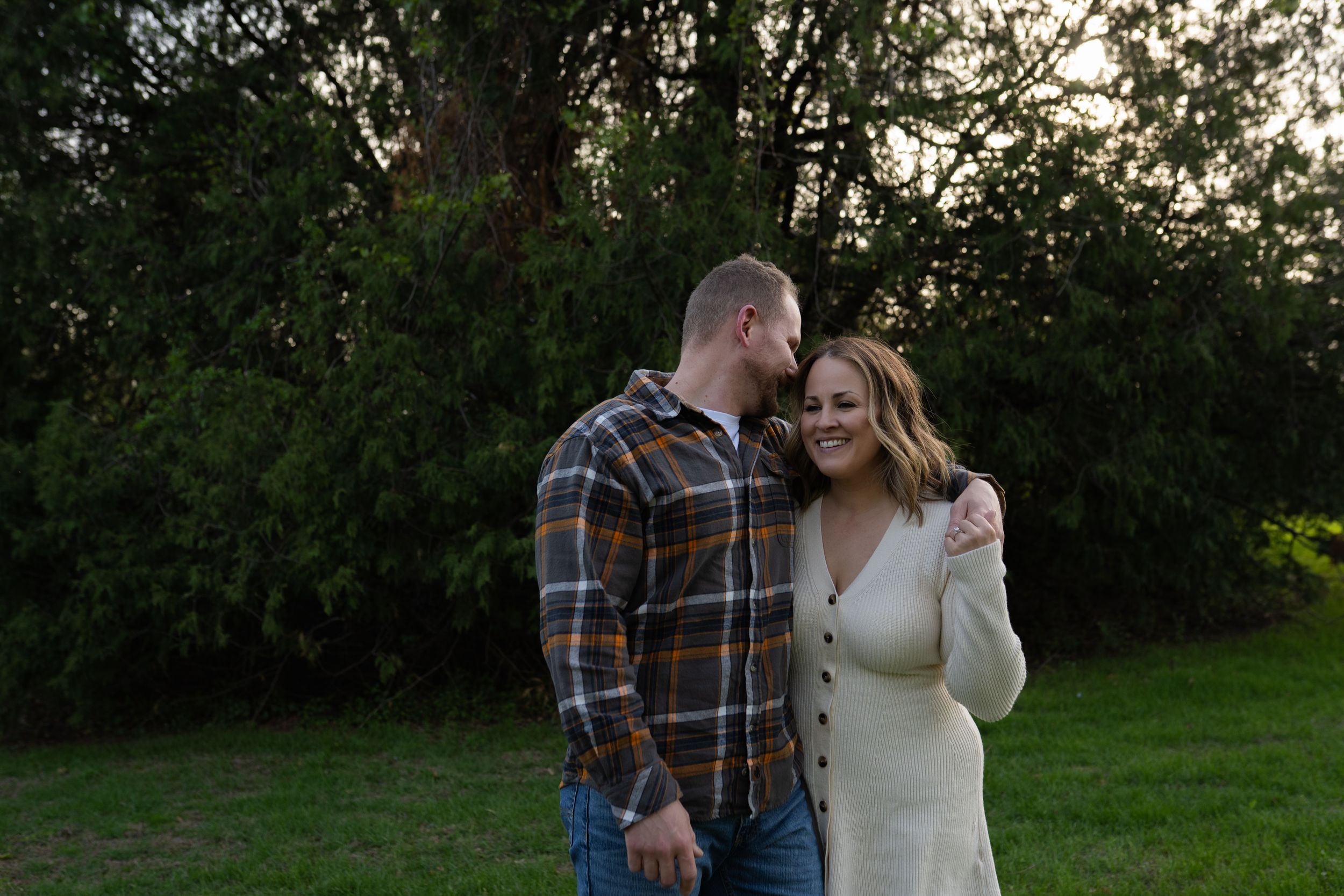 Couple smiling and embracing outdoors at dusk with lush green trees in background.