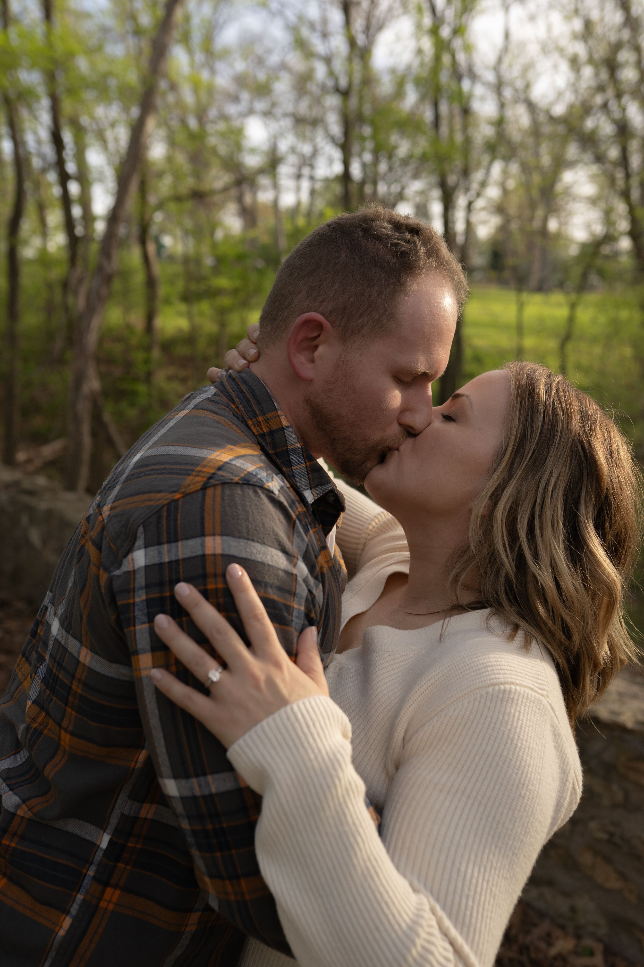 A couple shares a romantic kiss outdoors, surrounded by lush green trees in a serene forest setting.