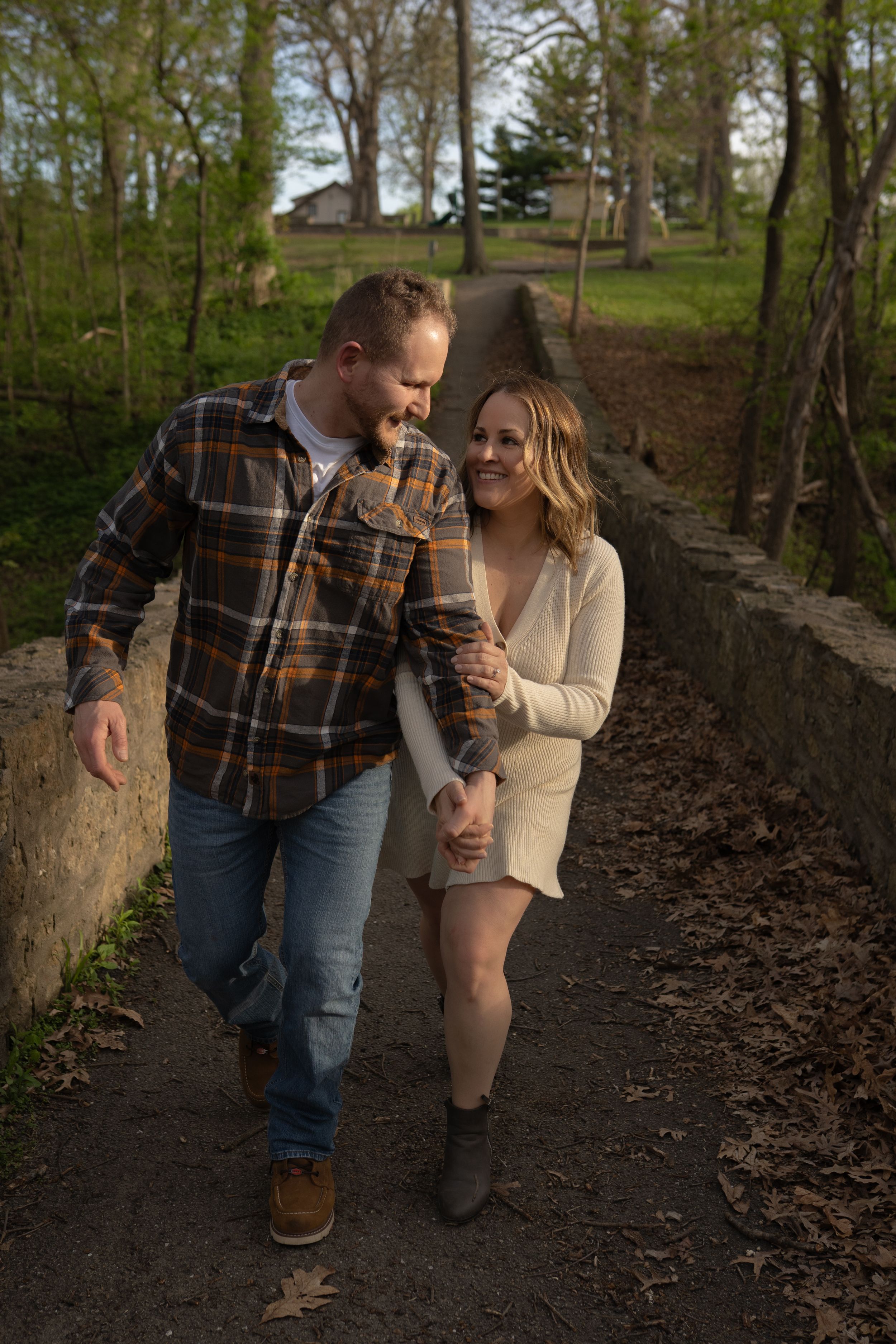 Couple walking hand-in-hand on a stone path in a park, smiling at each other, wearing casual fall outfits.