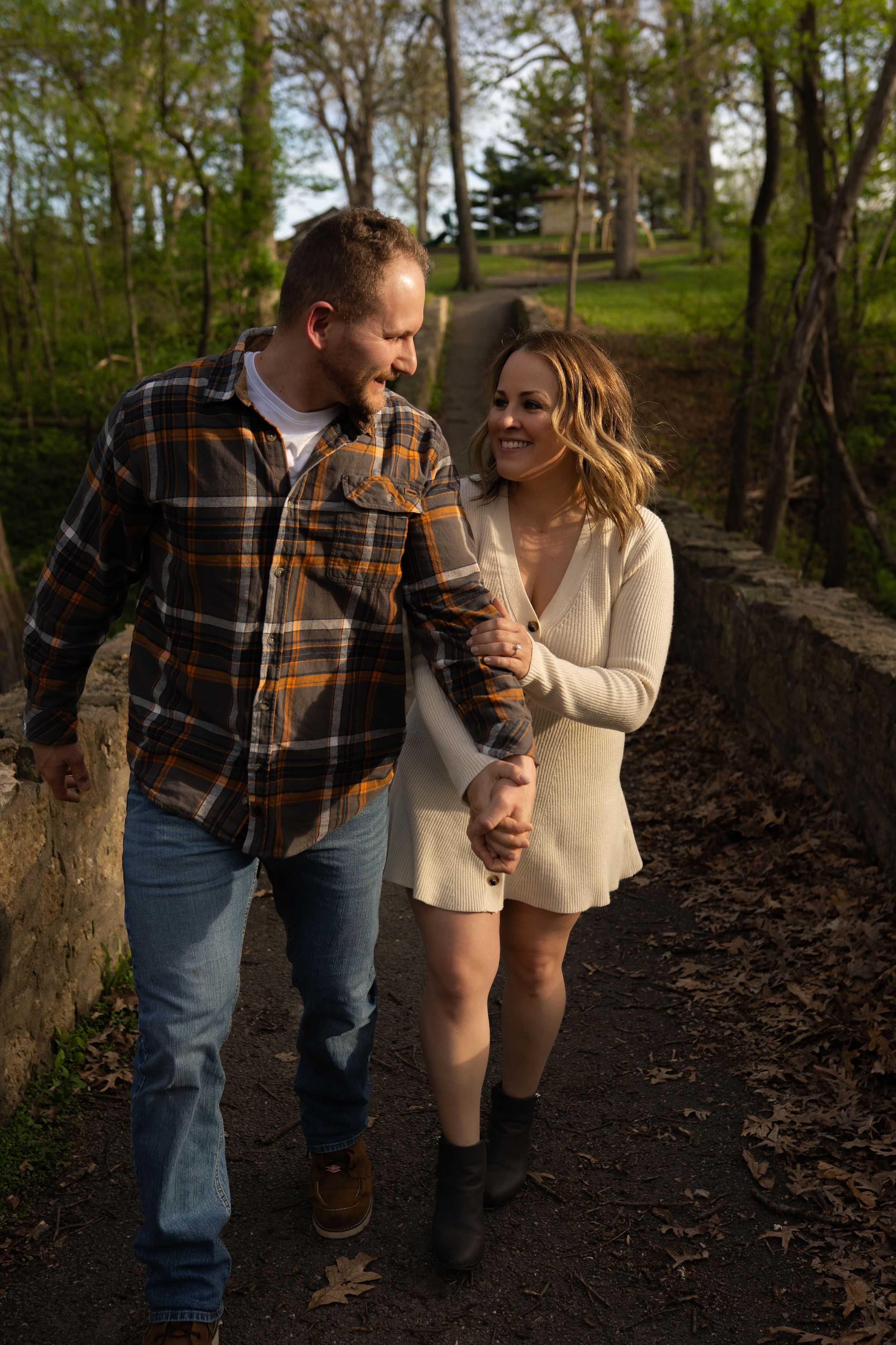 Couple holding hands and smiling while walking on a stone path surrounded by lush green trees.