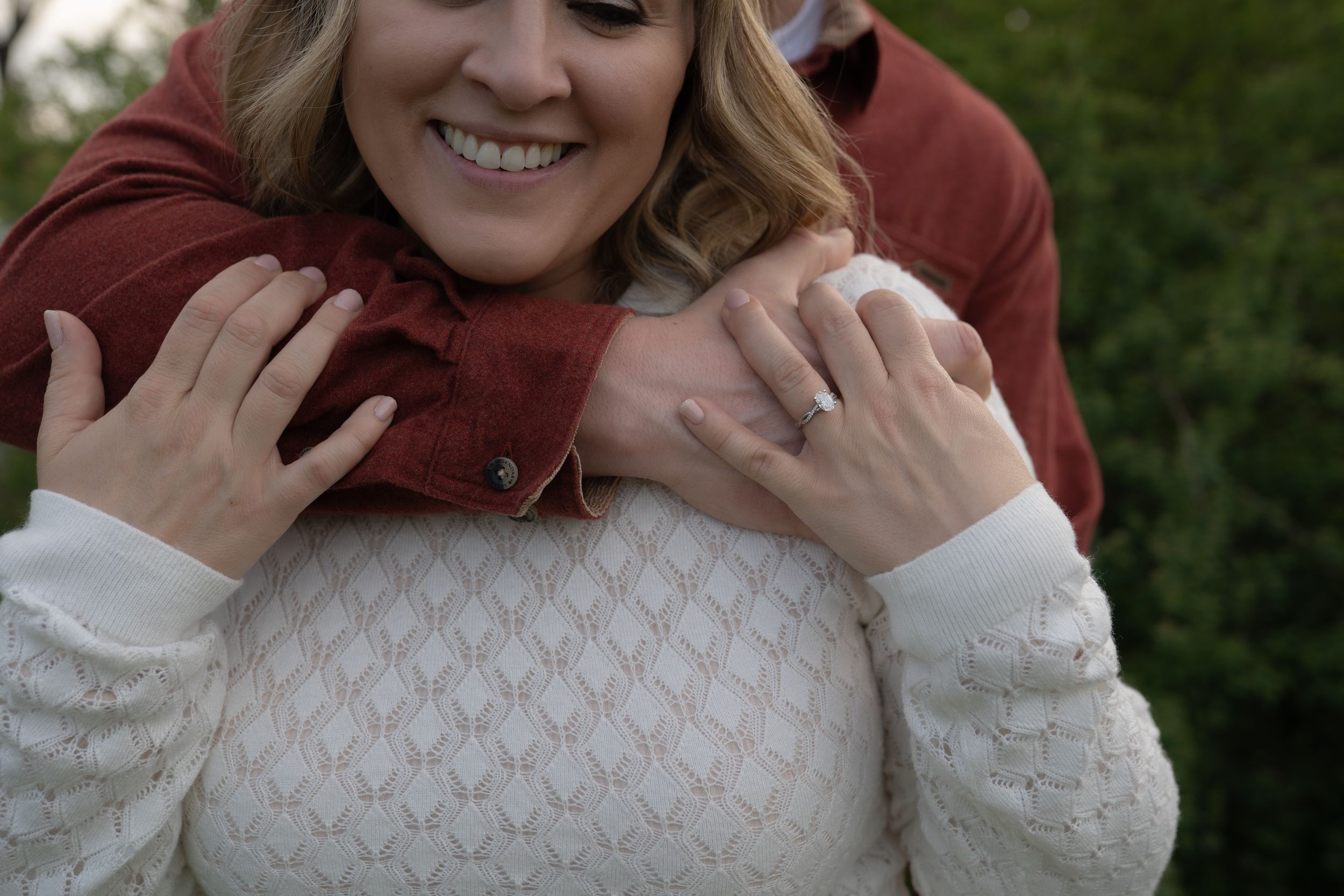 Couple embracing outdoors, woman in white lace sweater showing engagement ring, man in rust sweater hugging from behind.