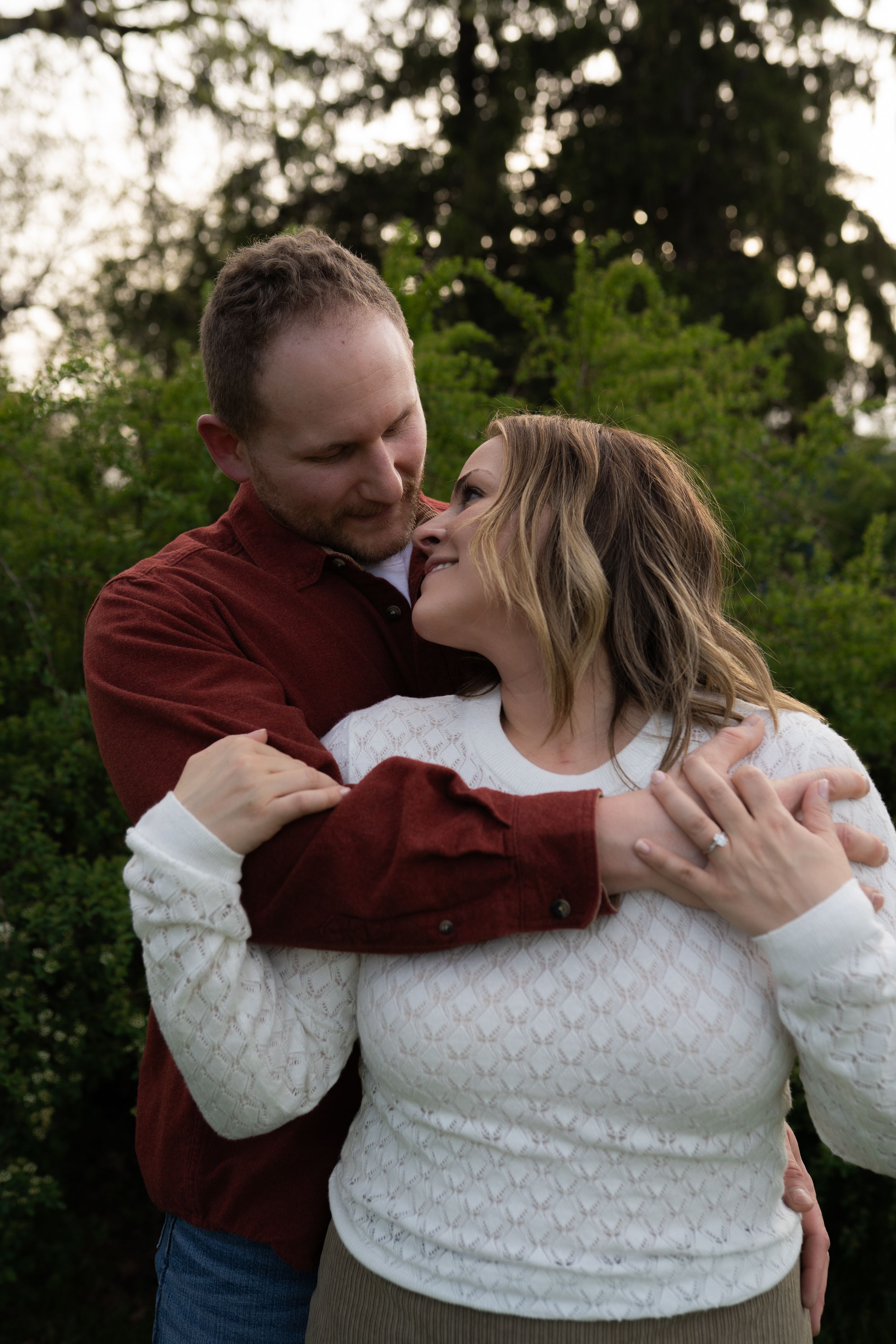 A couple shares a romantic embrace outdoors, the man in red hugging a smiling woman in white with green trees behind them.