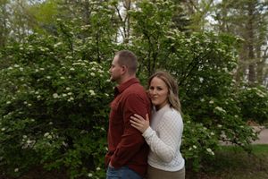 Engaged couple posing back-to-back in front of flowering white bushes in a lush garden setting.