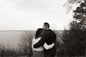 Black and white photo of a couple embracing overlooking a serene lake surrounded by bare trees.