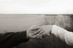 Black and white photo of couple holding hands, showing engagement ring, with blurred outdoor background.