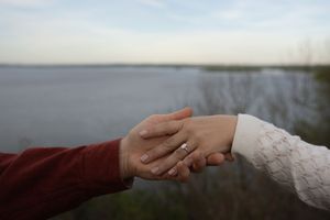 Close-up of couple holding hands showing engagement ring near a serene lakeside backdrop.
