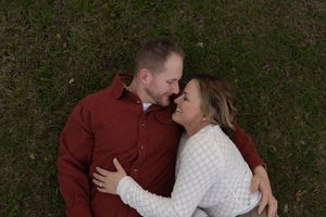 Engaged couple lying on green grass, smiling at each other in a romantic embrace.