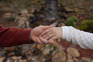 Couple holding hands outdoors showing engagement ring, rocky waterfall background.