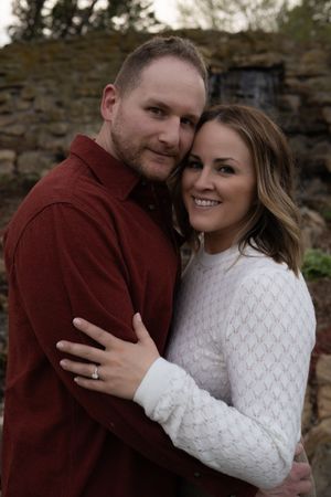 Happy couple hugging closely outdoors near a stone wall, woman wearing engagement ring.