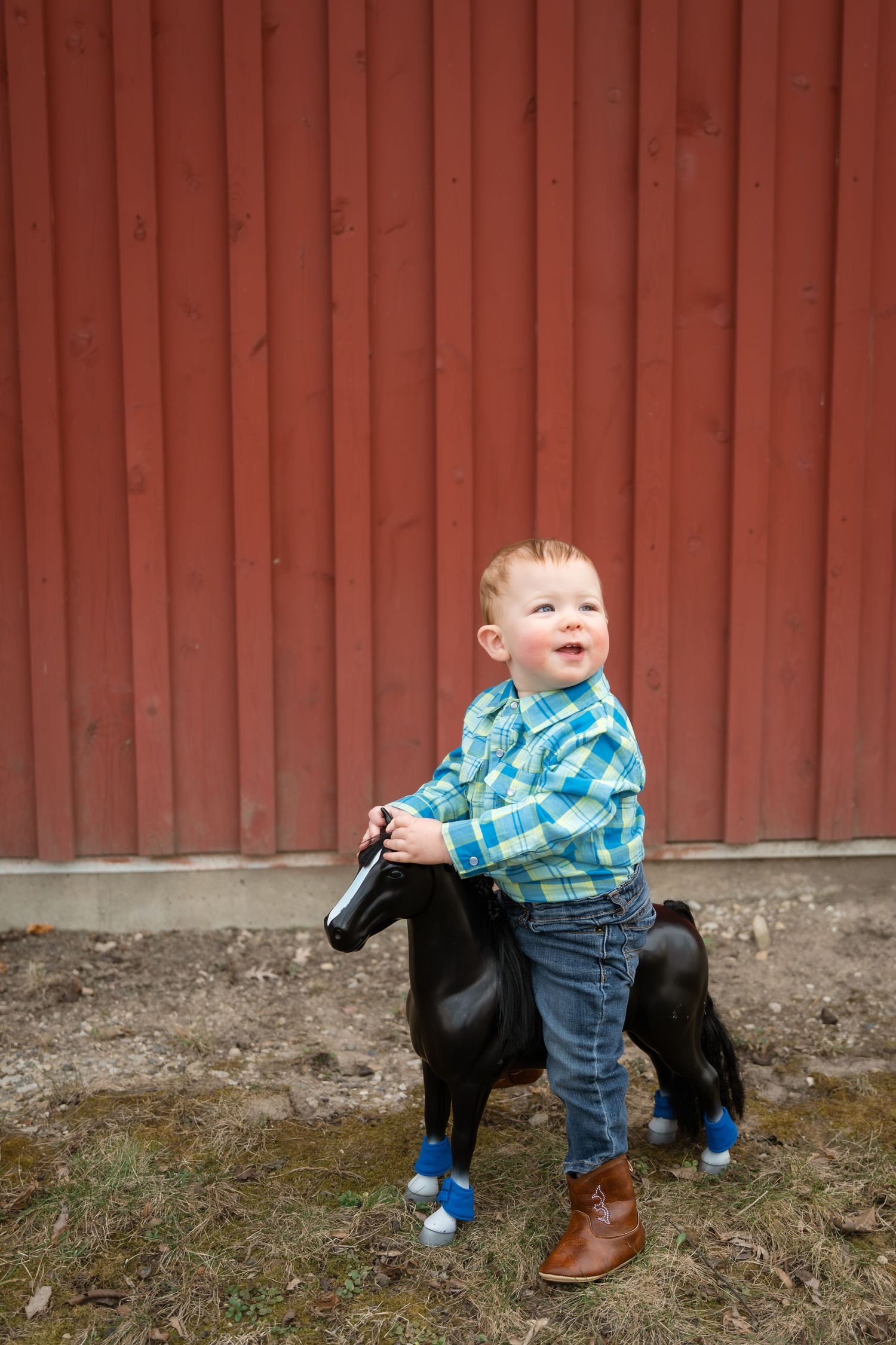 Smiling toddler boy in plaid shirt and jeans riding a black toy horse in front of a red barn wall.