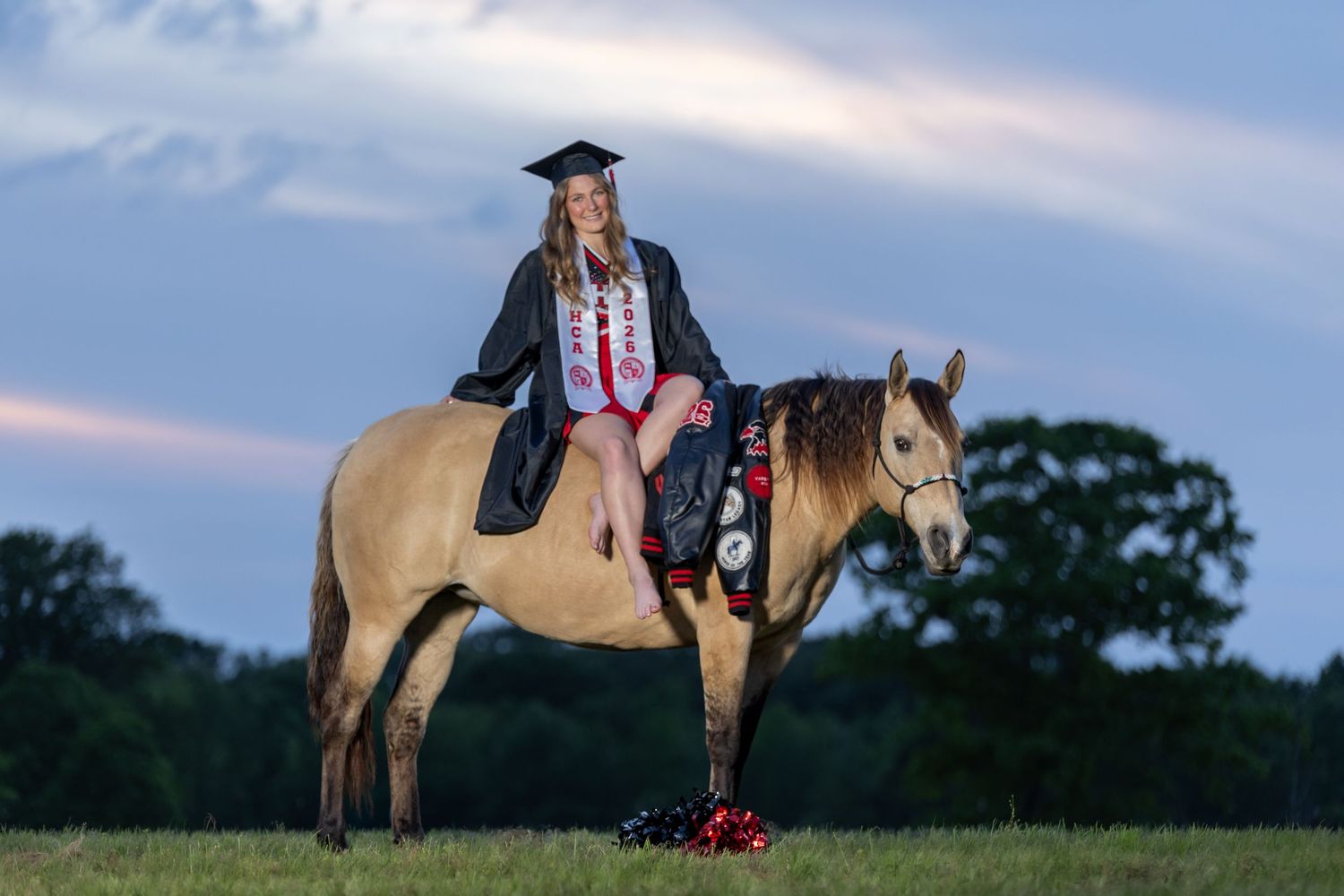 High school senior cheerleader equestrian senior portraits, girl sitting on horse wearing cheer uniform, cap and gown.