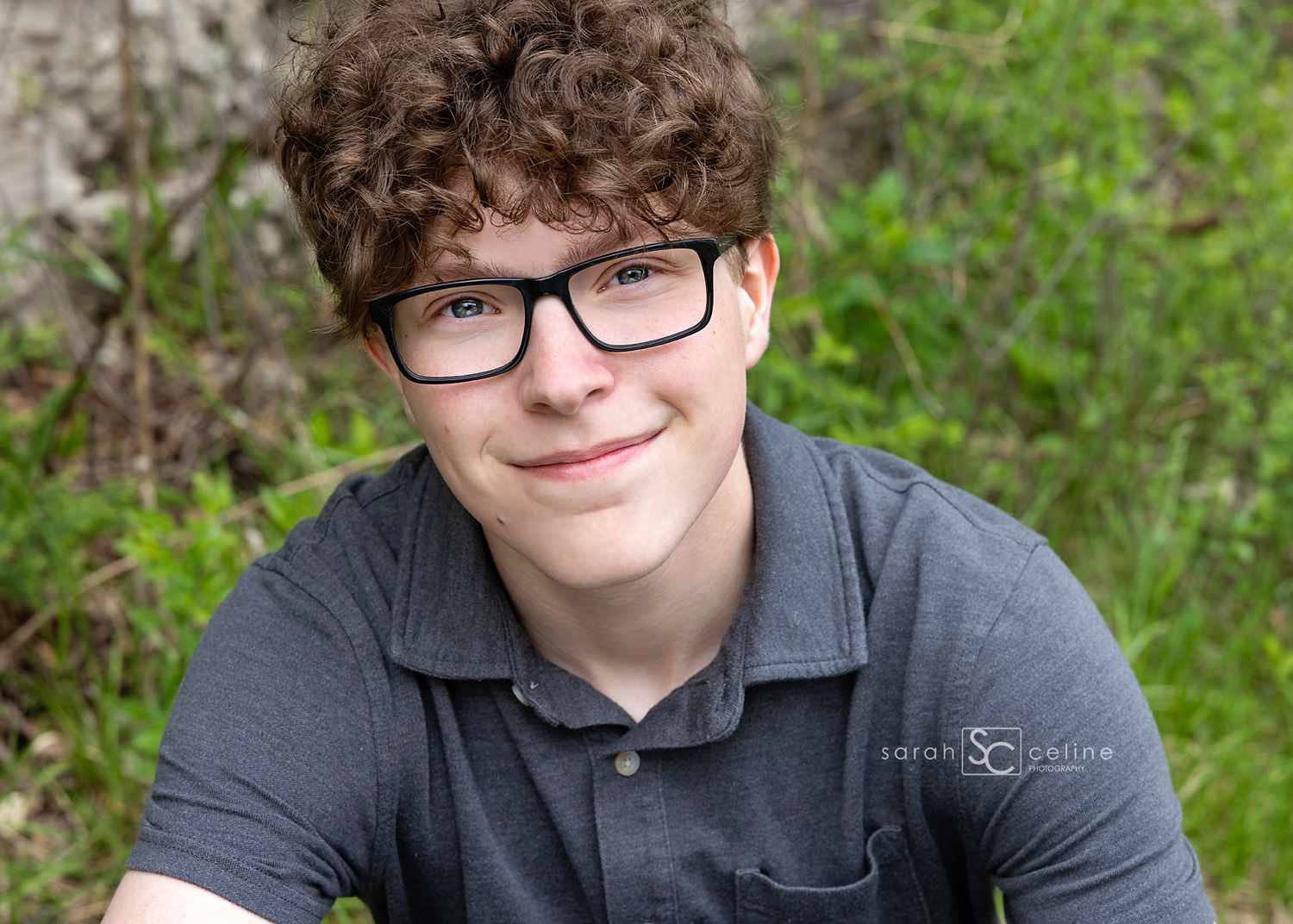 Teenage boy with curly brown hair and black glasses smiling outdoors in a green nature setting.