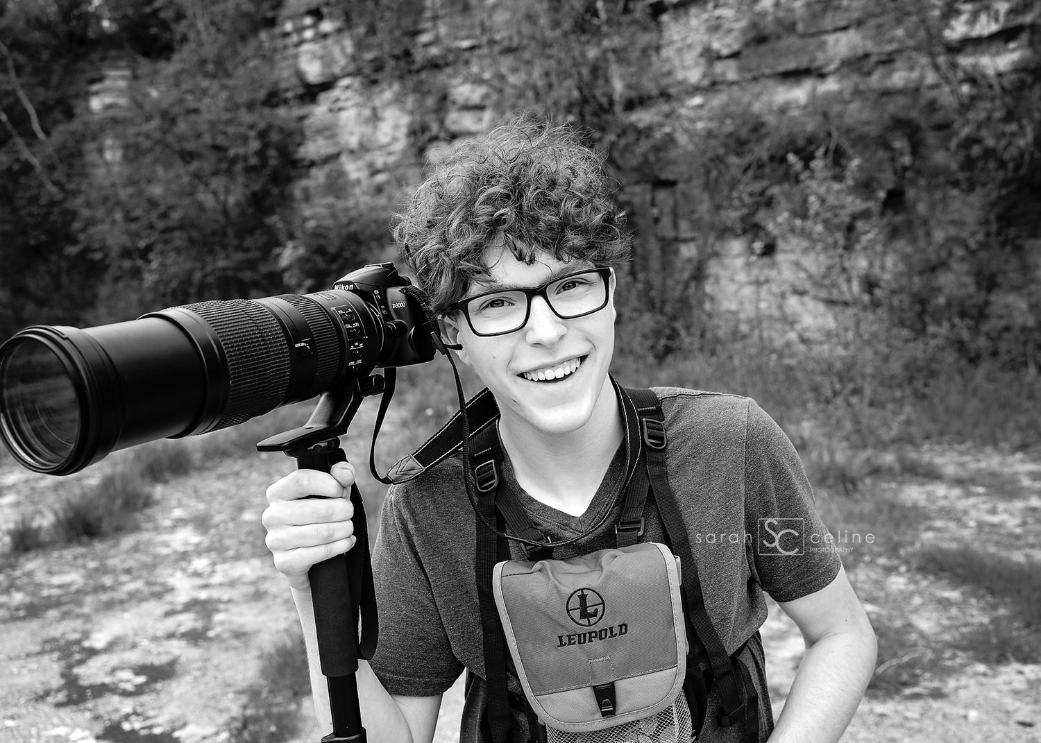 Young photographer smiling outdoors with large telephoto camera lens and Leupold binoculars in black and white.