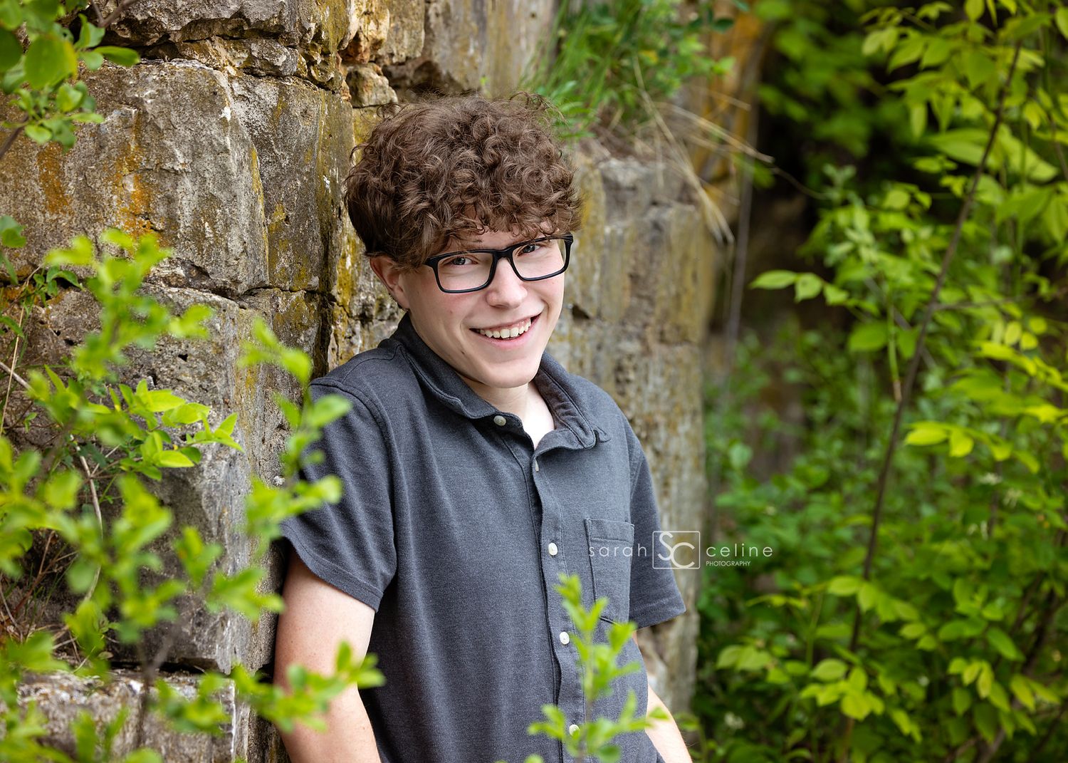 Smiling teenage boy with curly hair and glasses poses outdoors near a stone wall surrounded by green foliage.
