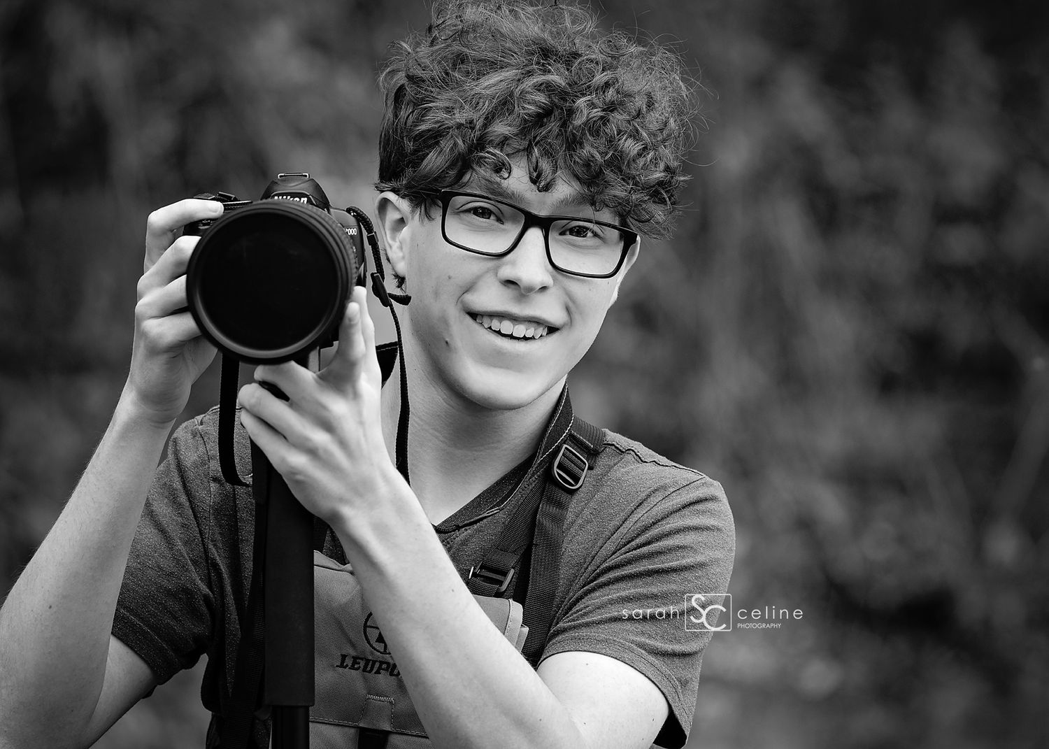 Young man with curly hair and glasses smiling while holding a professional DSLR camera outdoors.