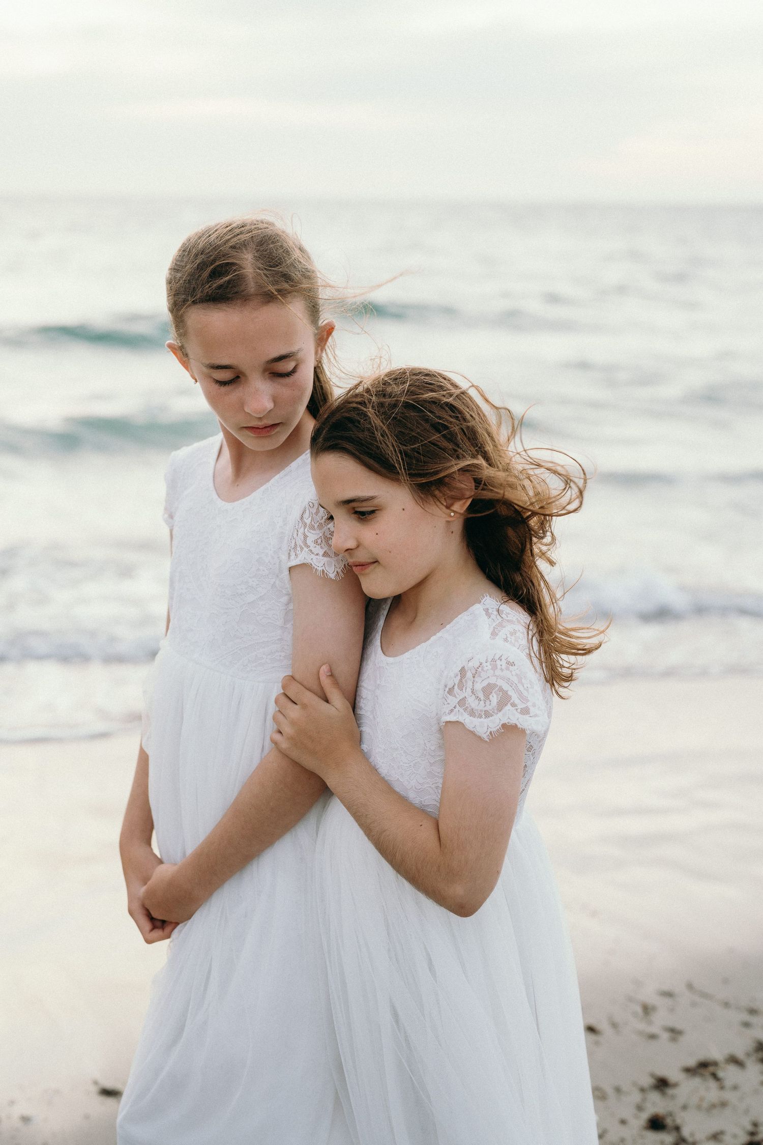 Two young girls in white lace dresses embracing on a beach with ocean waves in the background.