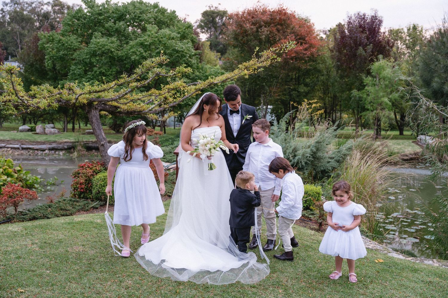 Bride and groom surrounded by flower girls and ring bearers in a lush garden setting during an outdoor wedding ceremony.