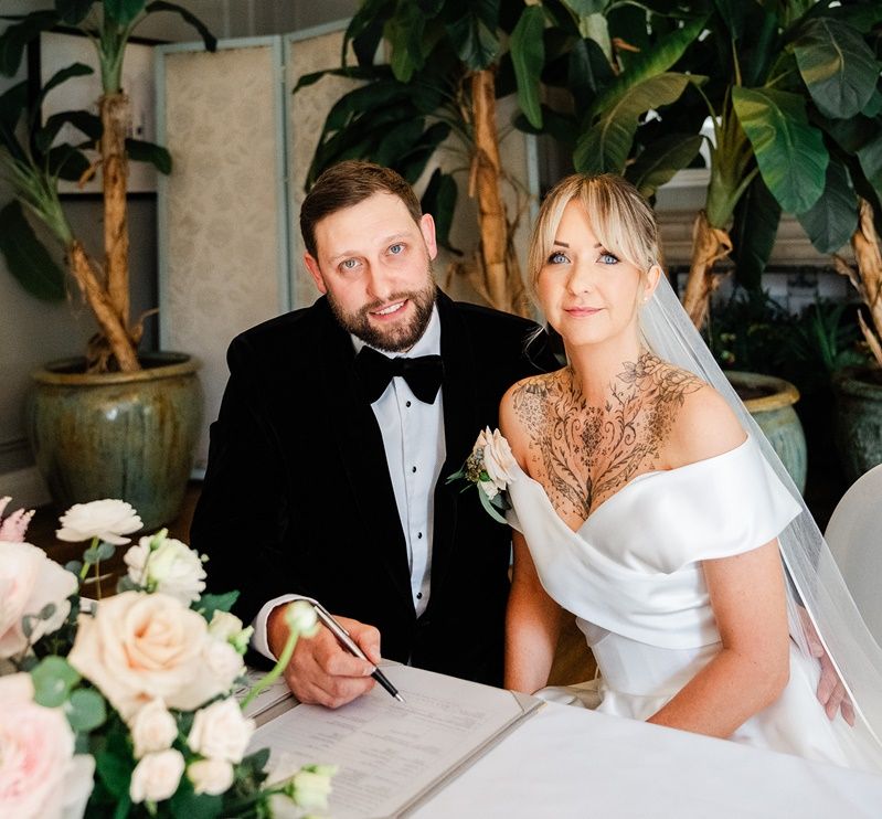 Bride and groom signing wedding registry, seated at table with pink floral arrangement, groom in tuxedo, bride in off-shoulder gown.
