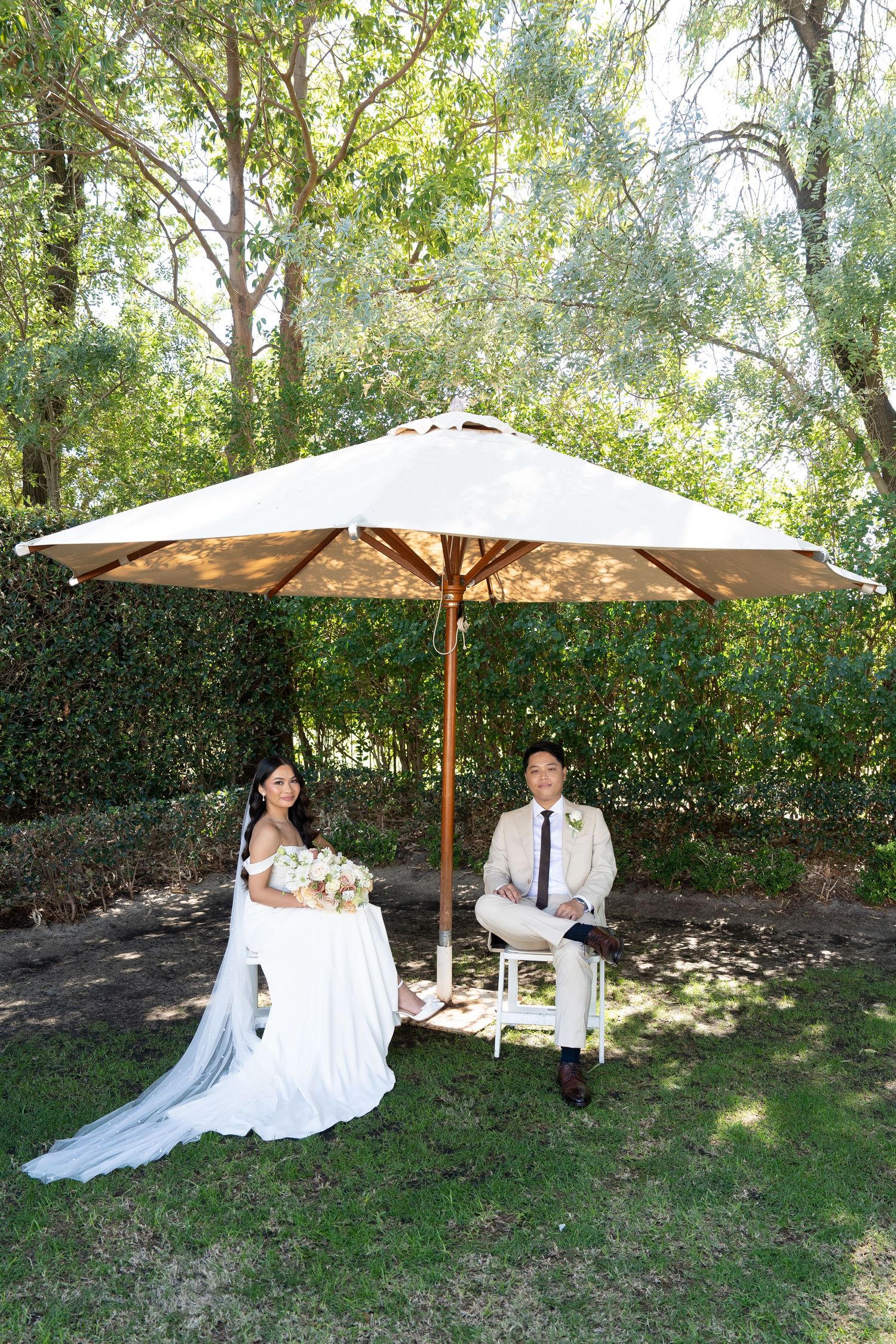 Bride and groom sit under a large patio umbrella in a lush garden setting during their outdoor wedding celebration.