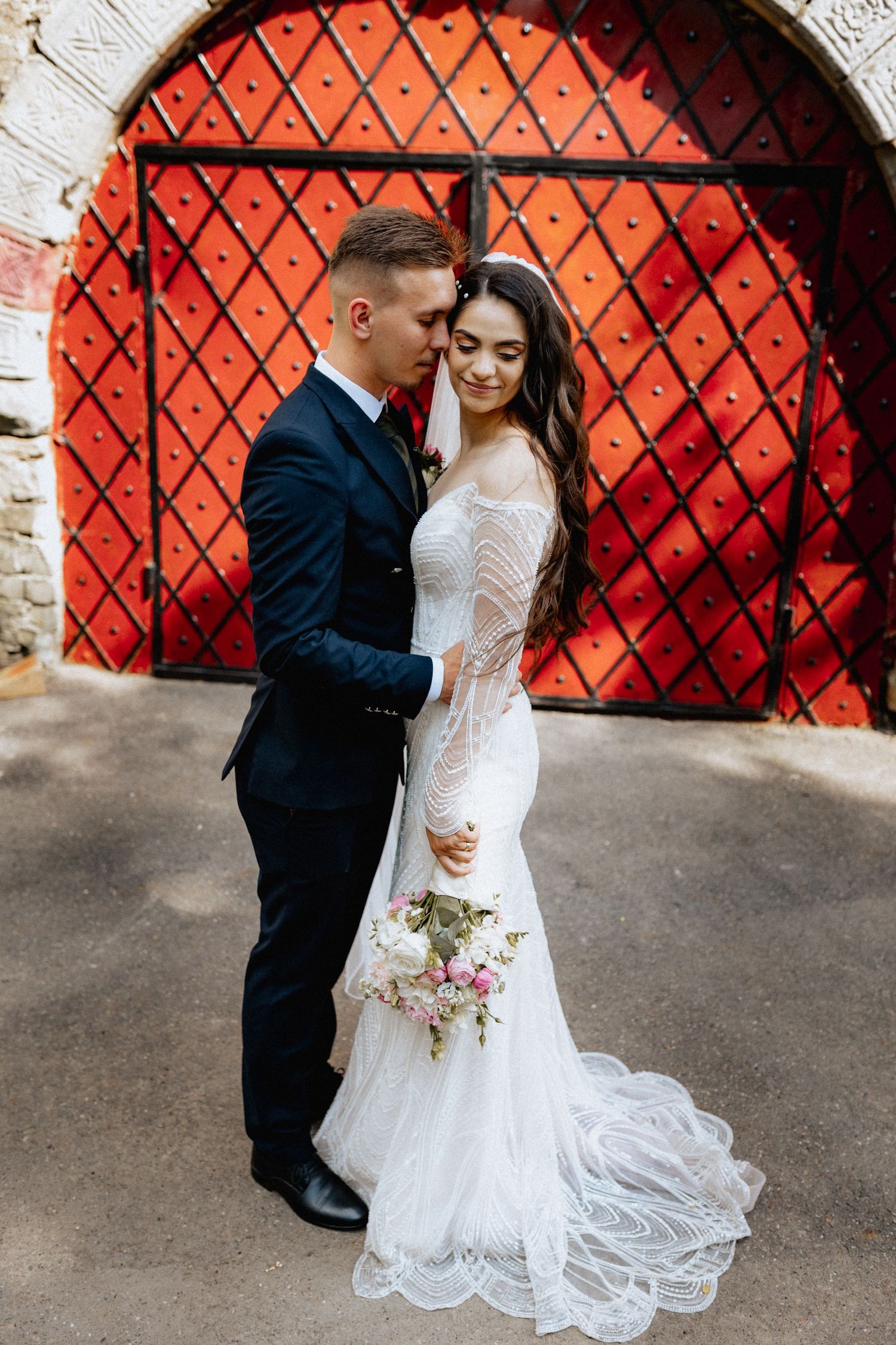 Bride and groom embracing in front of a red diamond-patterned door, bride holding a pink bouquet in a white lace gown.
