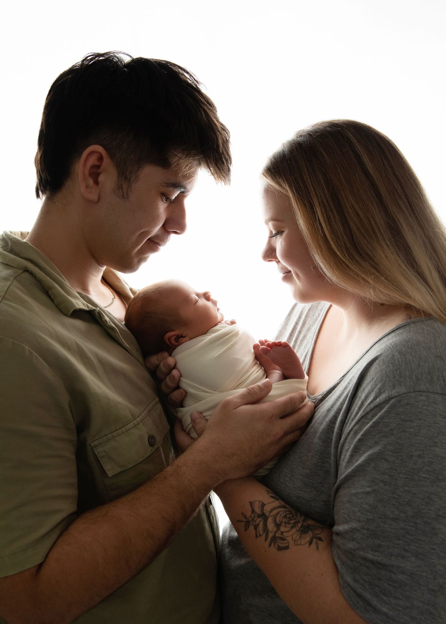 New parents tenderly hold their swaddled newborn baby together, sharing a loving moment in soft backlit light.