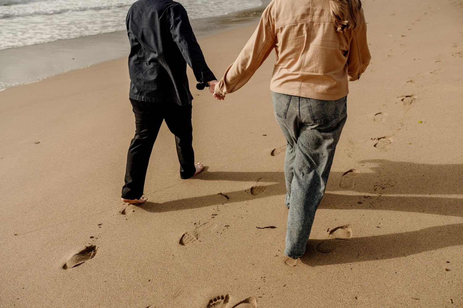 Two people holding hands walking barefoot on a sandy beach, leaving footprints in the sand near the ocean.