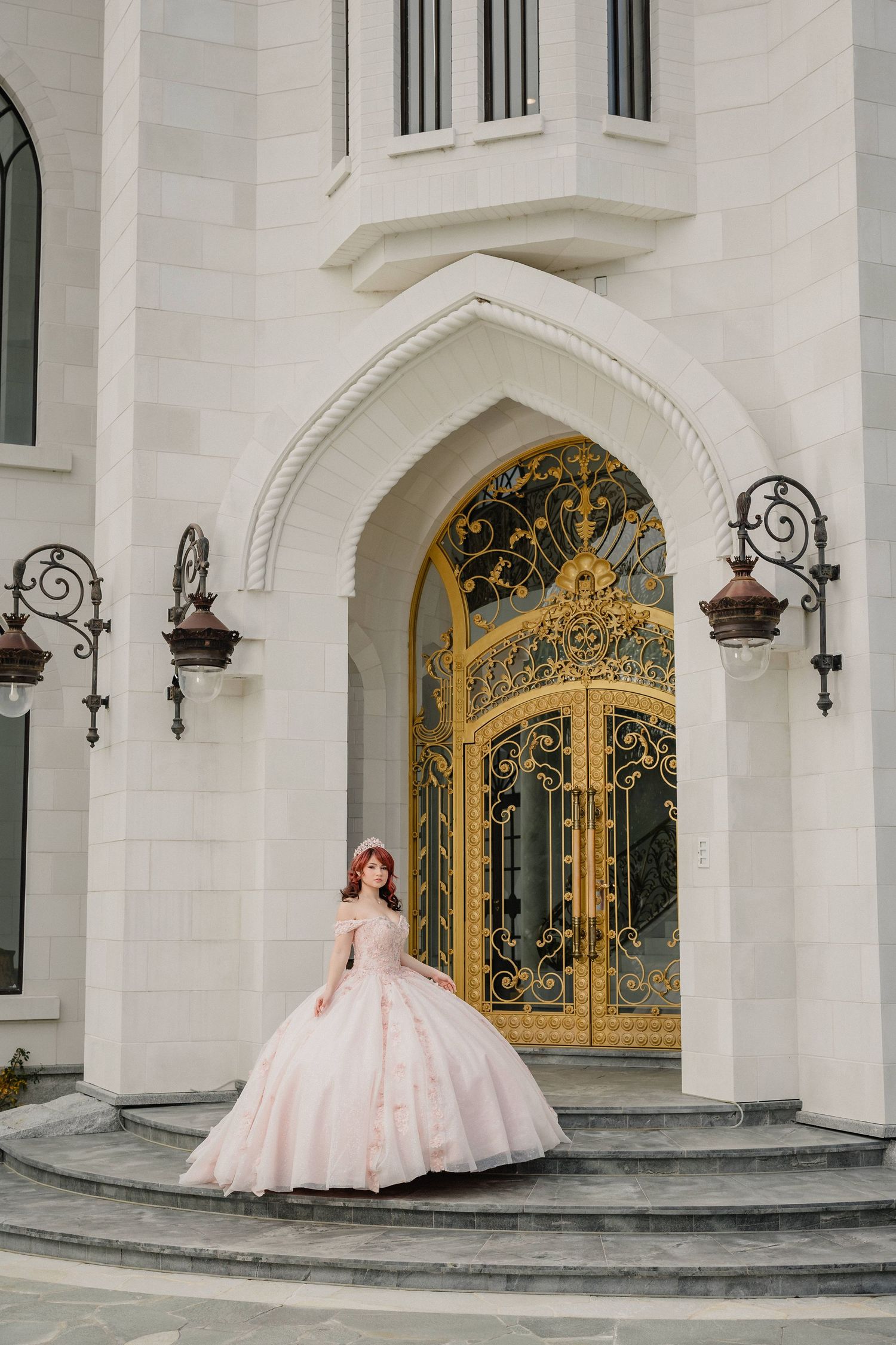 Woman in pink ballgown poses on steps before ornate golden gates of white castle-like building.