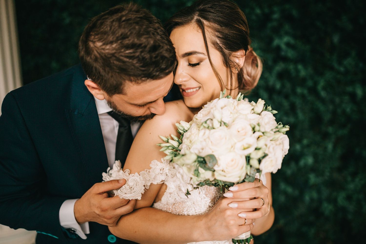 A groom in a navy suit embraces his smiling bride holding a white floral bouquet on their wedding day.