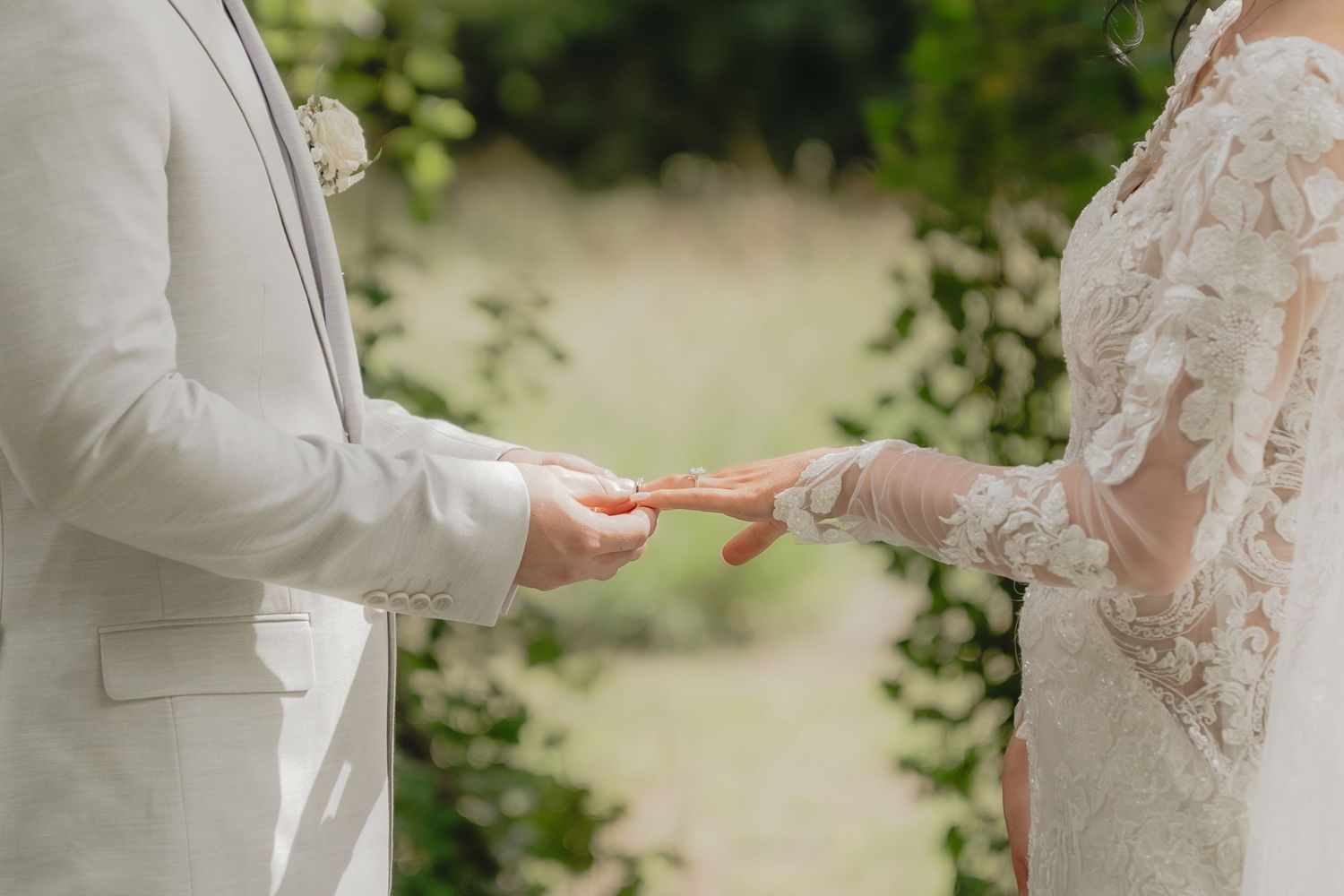 Bride and groom exchanging rings during outdoor wedding ceremony, bride wearing lace gloves and gown.