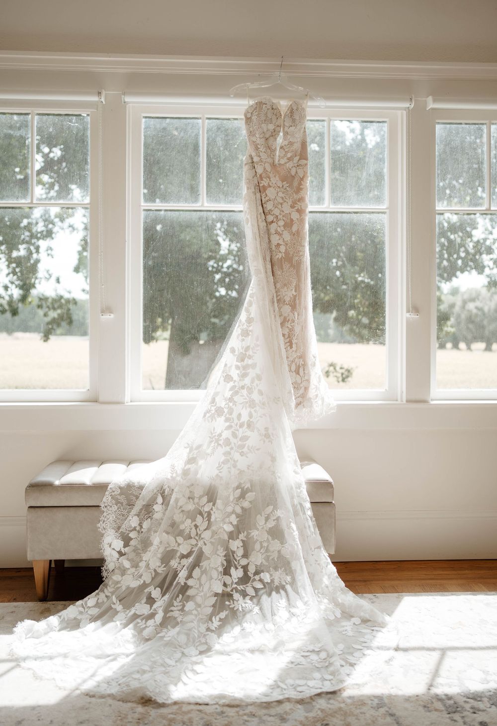 Elegant lace wedding dress with floral appliques and long train hanging by sunlit windows.