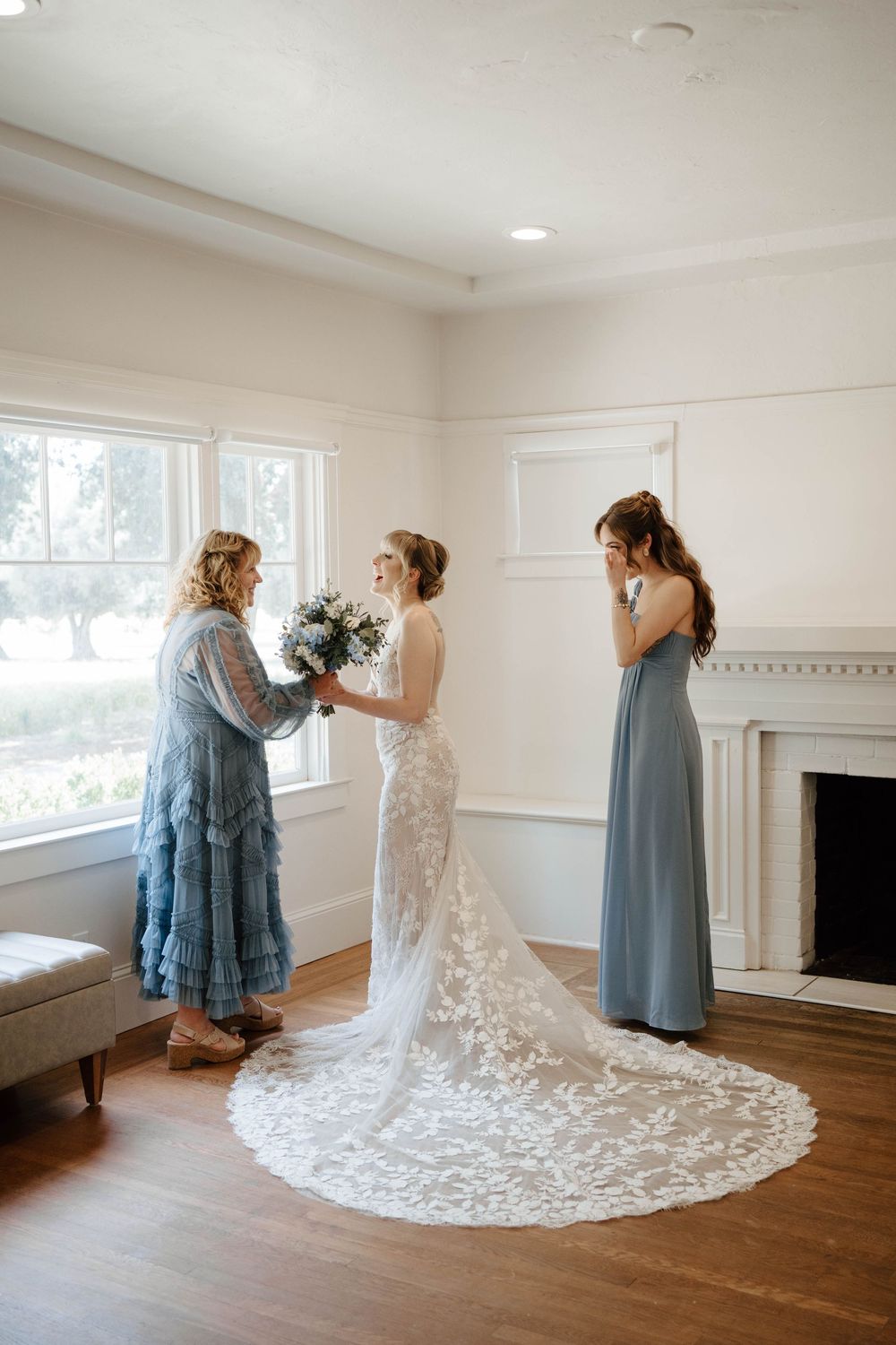 Bride in lace gown receives bouquet from woman in blue dress as bridesmaid looks on near fireplace.