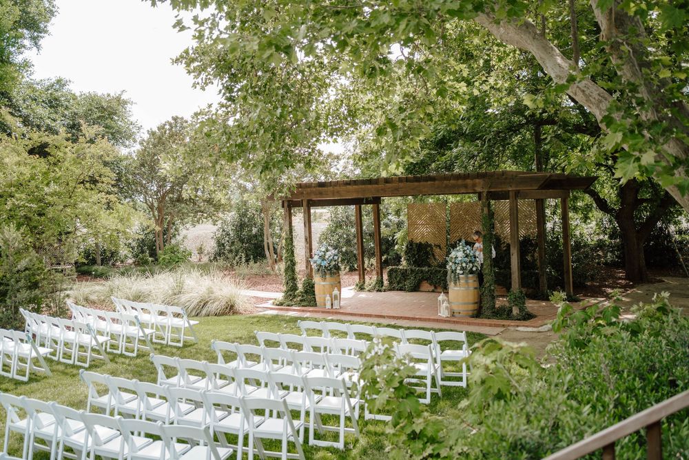 Outdoor wedding ceremony setup with white chairs on green lawn facing rustic wooden pergola altar surrounded by lush garden trees.