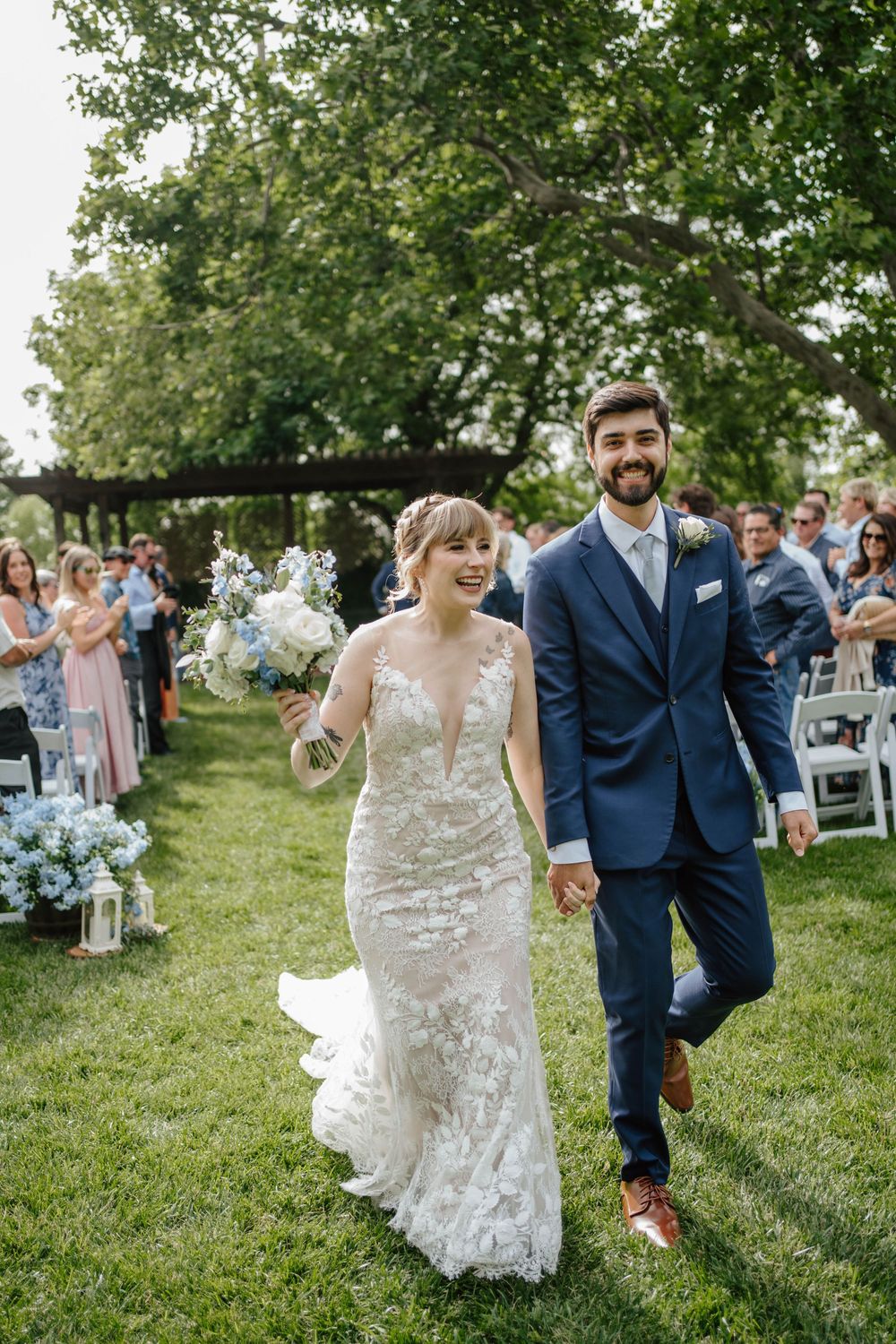 Smiling bride in lace gown and groom in navy suit walk hand-in-hand down outdoor wedding aisle lined with guests.