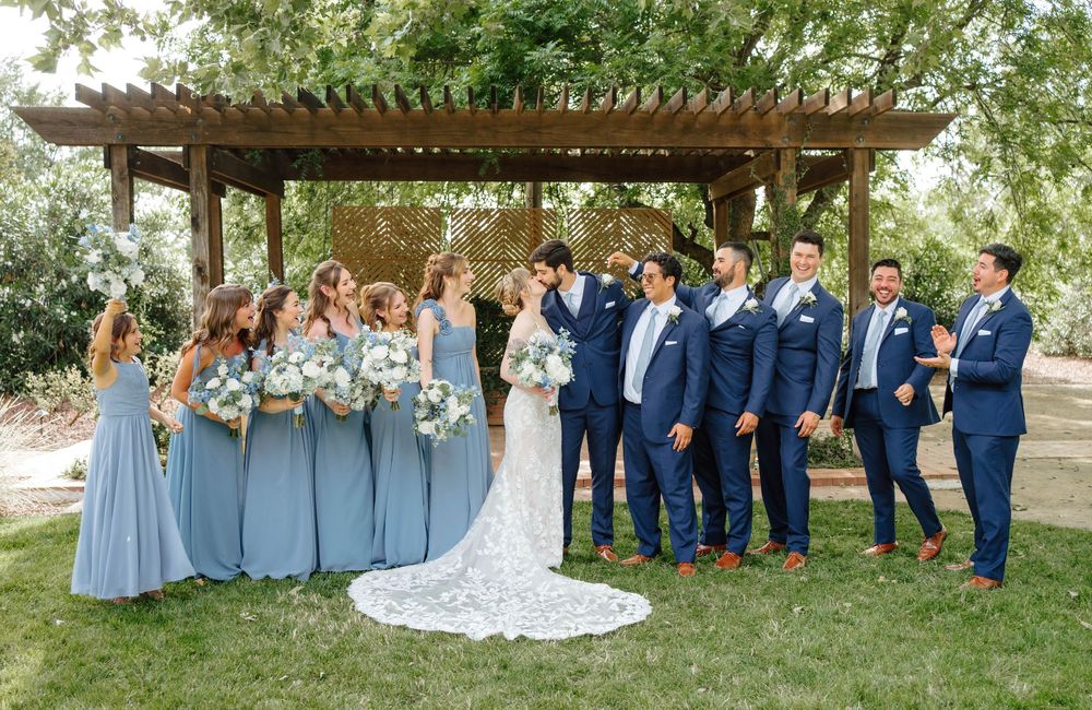Wedding party in navy suits and blue bridesmaid dresses celebrating outdoors under a wooden pergola.