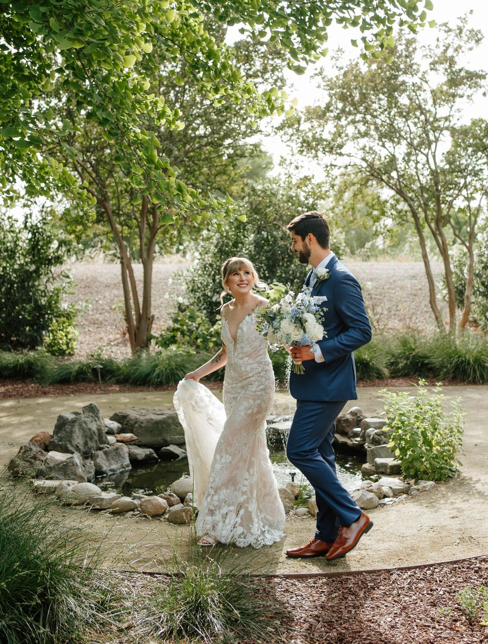 Bride in lace gown and groom in navy suit walk through a lush garden, holding a white and blue bouquet.