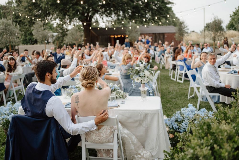 Bride and groom seated at outdoor wedding reception table with guests raising hands in celebration under string lights.