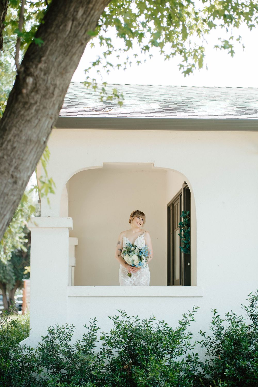 Elegant bride in lace gown holding blue and white bouquet standing in arched doorway of white building.