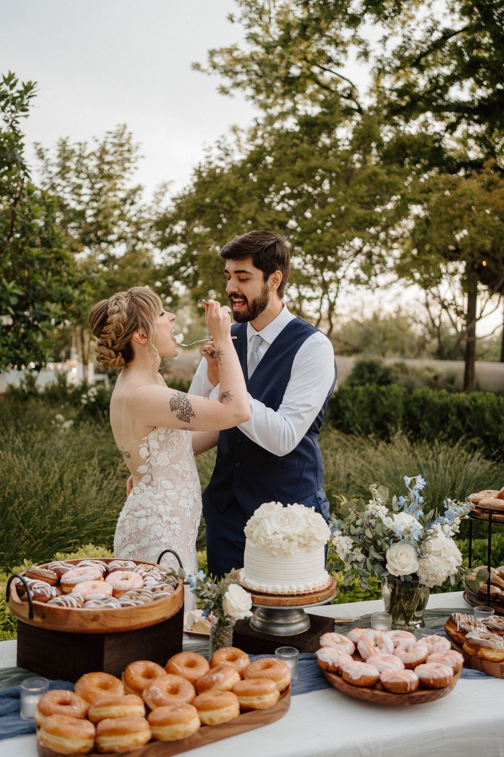 Bride and groom share cake at outdoor wedding dessert table filled with donuts and a white floral cake.