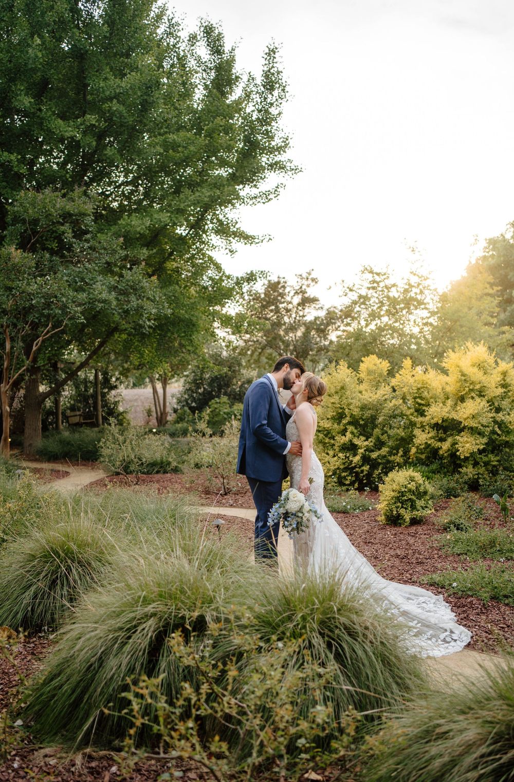 Bride and groom share romantic kiss in lush garden at golden hour during outdoor wedding ceremony.