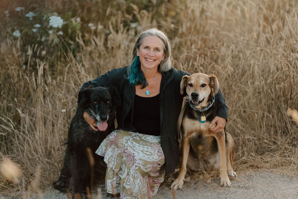 Woman smiling outdoors with two dogs, a black dog and a tan dog, sitting on a dirt path among golden grasses.