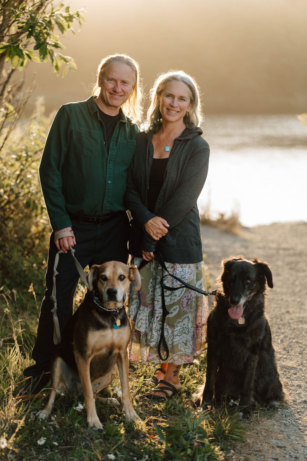 Smiling couple with two dogs on a scenic outdoor path near water at golden hour.