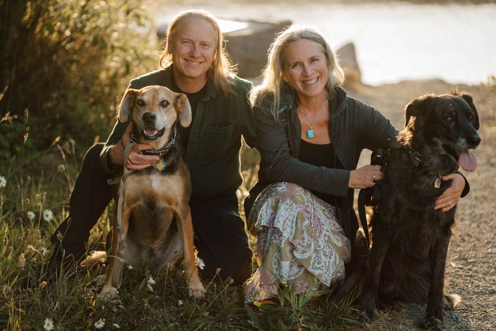 Two smiling people pose outdoors with a beagle and black Labrador near a lake at golden hour.